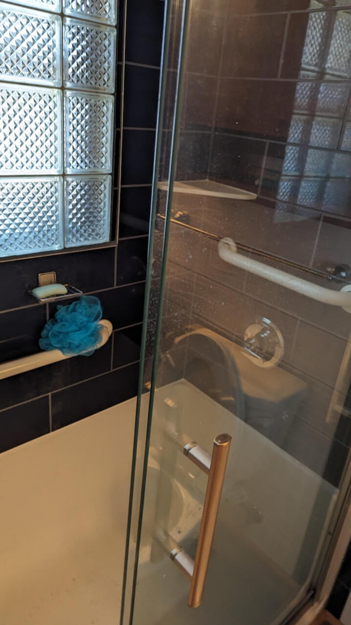 Glass shower door with a metal handle, a blue bath pouf hanging on a grab bar, and tiled walls surrounding a bathtub—expertly installed by Third Day Plumbing - Des Moines Iowa.
