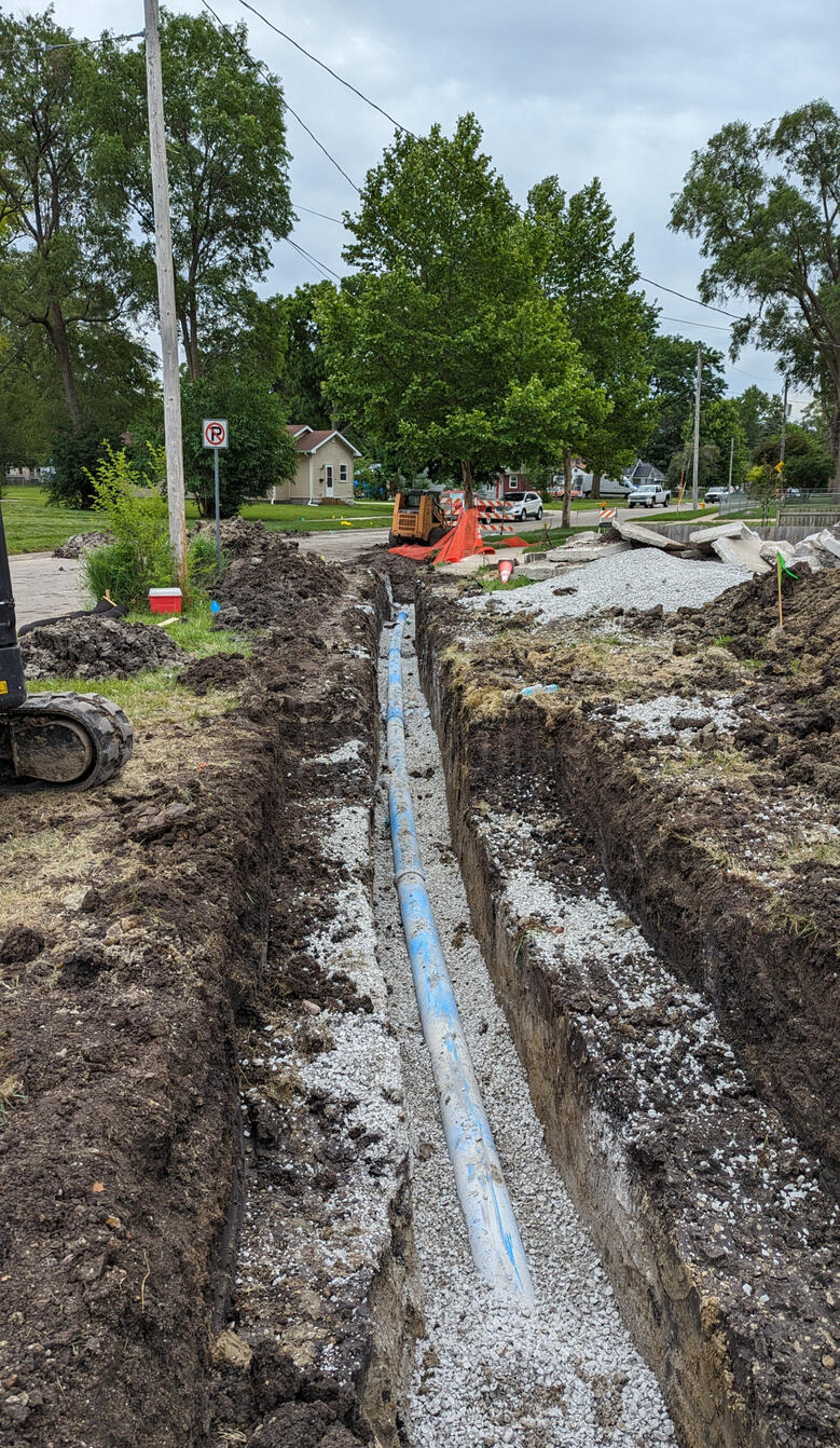 A blue water pipe lies in a freshly dug trench along a roadside construction site, with gravel and construction equipment visible in the background.